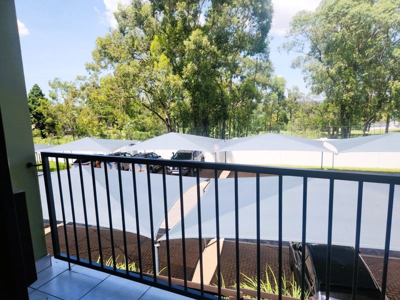 A balcony with a metal balustrade overlooking a paved parking area with shade sail structures and surrounding trees.