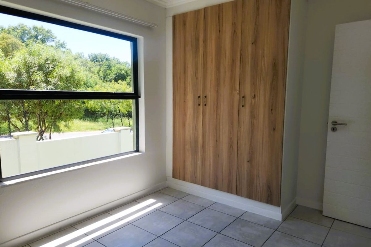 A bright, empty bedroom featuring modern wood-finish built-in cupboards, neutral tiled flooring, and a large window with a pleasant outdoor view.