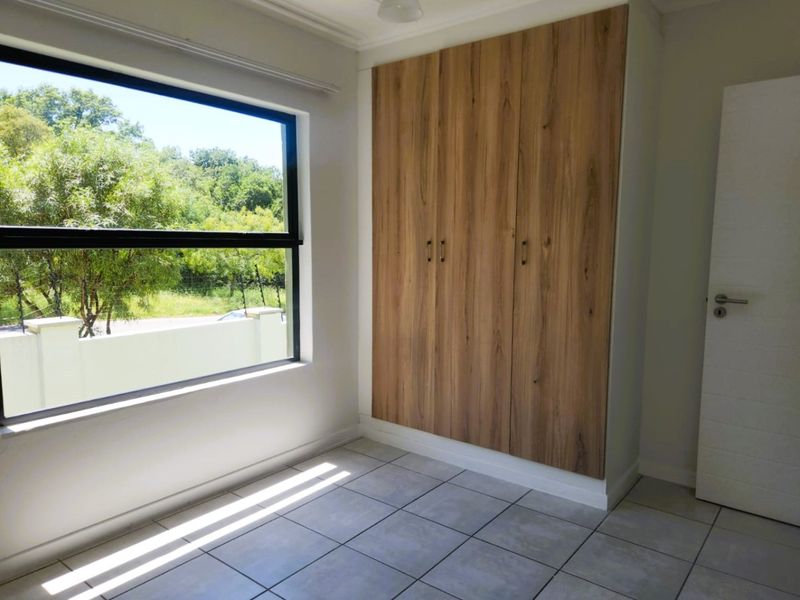 A bright, empty bedroom featuring modern wood-finish built-in cupboards, neutral tiled flooring, and a large window with a pleasant outdoor view.