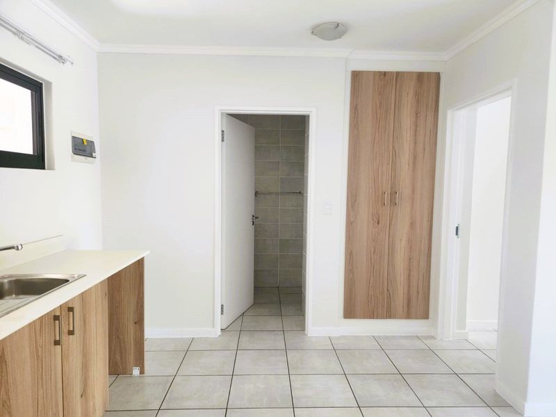 A clean, modern kitchen area featuring light wood-finish cabinetry, tiled flooring, and a built-in cupboard, leading into an adjacent tiled bathroom space.
