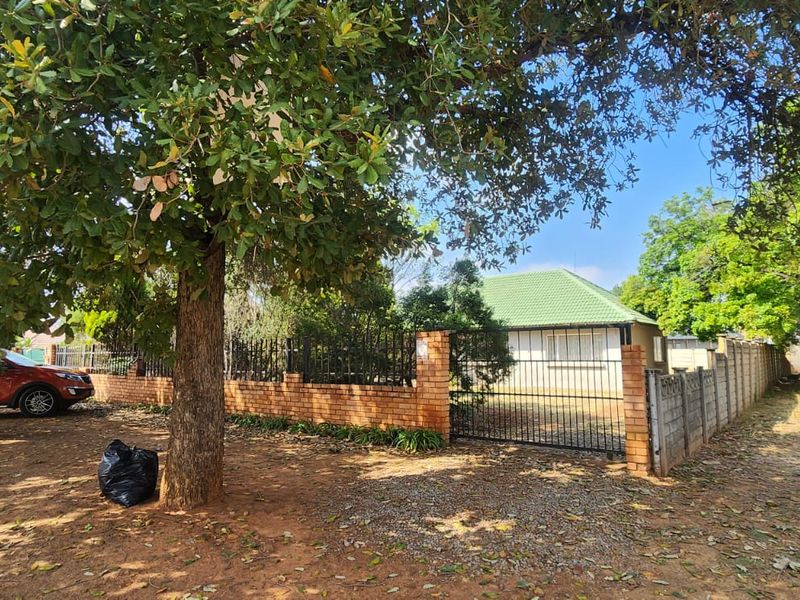 Front view of a residential property featuring a brick boundary wall, metal security gate, and a tiled roof structure surrounded by mature trees.