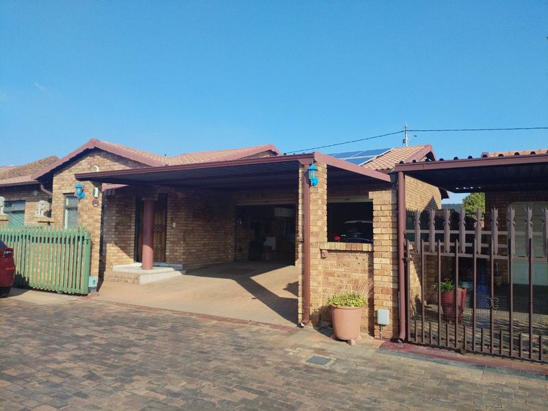 Front exterior view of a face brick townhouse featuring a covered carport, paved driveway, solar panels, and secure metal gate access.