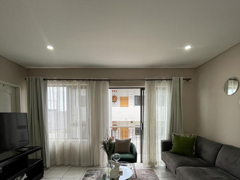 A well-lit living room featuring tiled flooring, recessed downlights, and a sliding glass door providing access to the balcony.