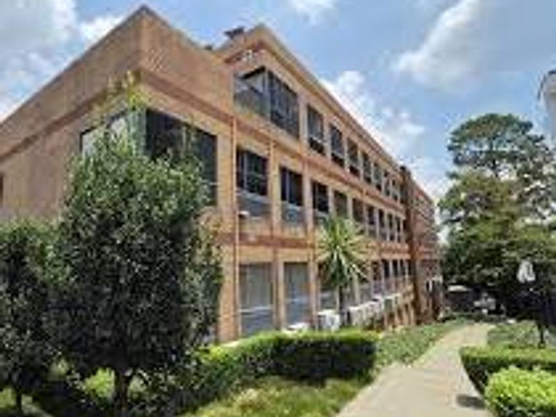 A multi-story commercial brick building featuring large windows, external air conditioning units, and a well-maintained paved walkway surrounded by landscaped garden areas.
