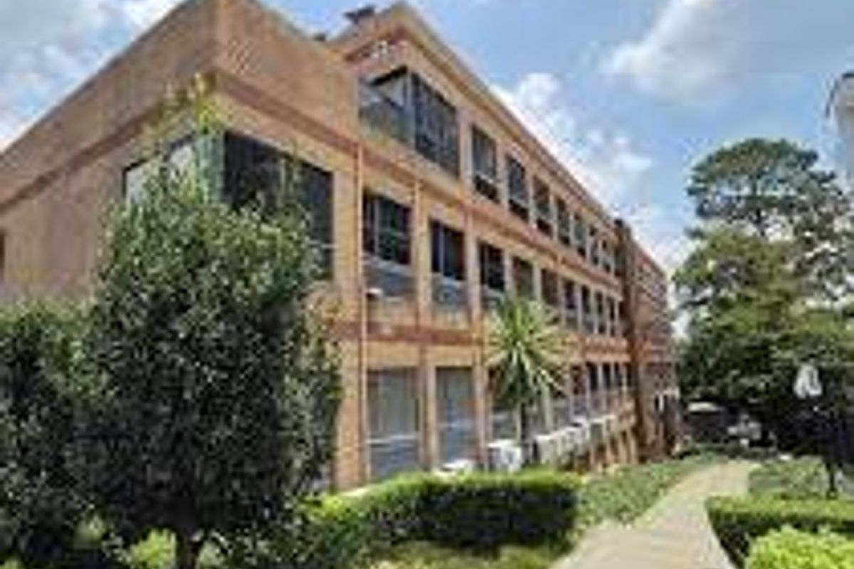 A multi-story commercial brick building featuring large windows, external air conditioning units, and a well-maintained paved walkway surrounded by landscaped garden areas.