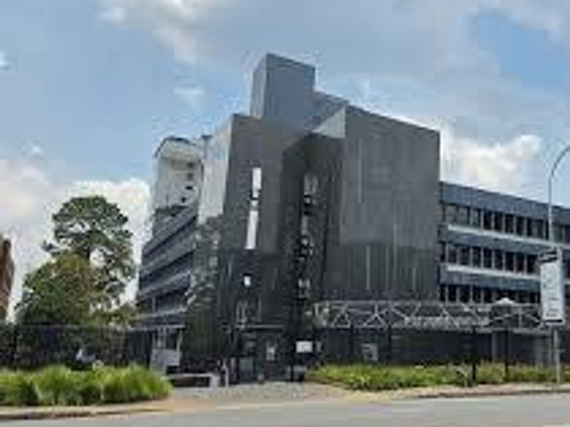 A modern multi-story commercial building featuring dark cladding, secure perimeter fencing, and a paved approach from the street.