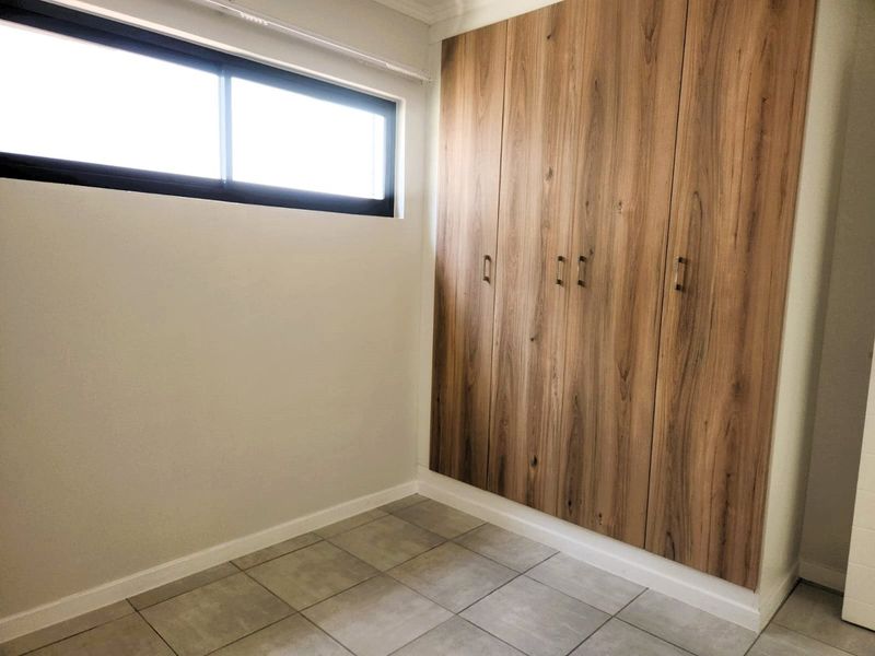 A clean, empty bedroom featuring modern wood-finish built-in cupboards, neutral tiled flooring, and a large sliding window providing natural light.