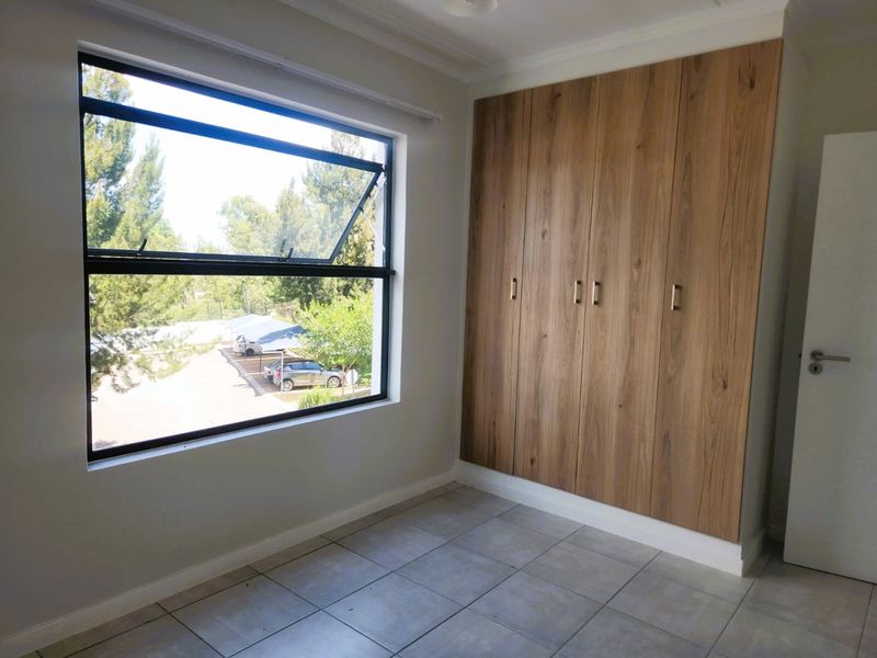 A bright, empty bedroom featuring modern wood-finish built-in cupboards, neutral tiled flooring, and a large window with views of the complex grounds.