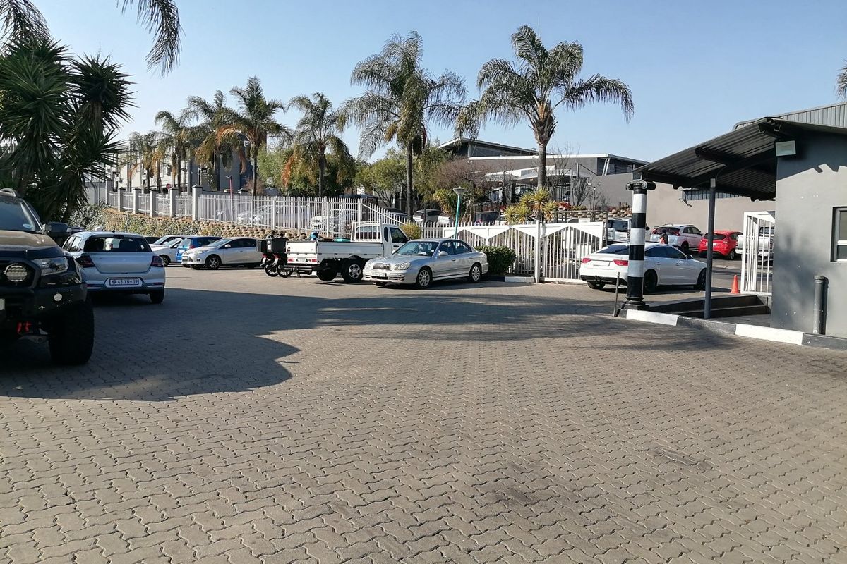 A paved commercial parking area and driveway entrance featuring security fencing, mature palm trees, and a covered entrance canopy.