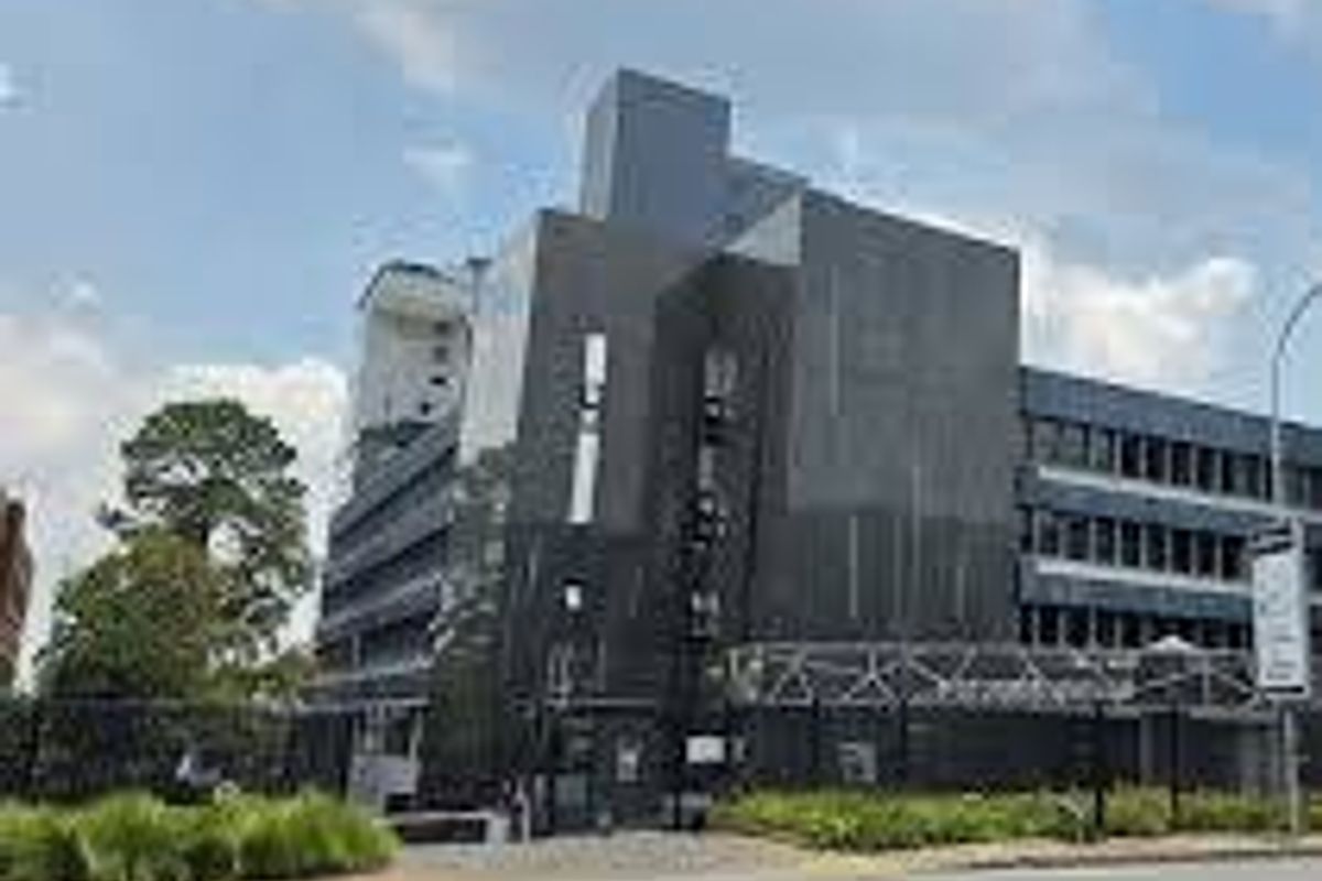 A modern multi-story commercial building featuring dark cladding, secure perimeter fencing, and a paved approach from the street.