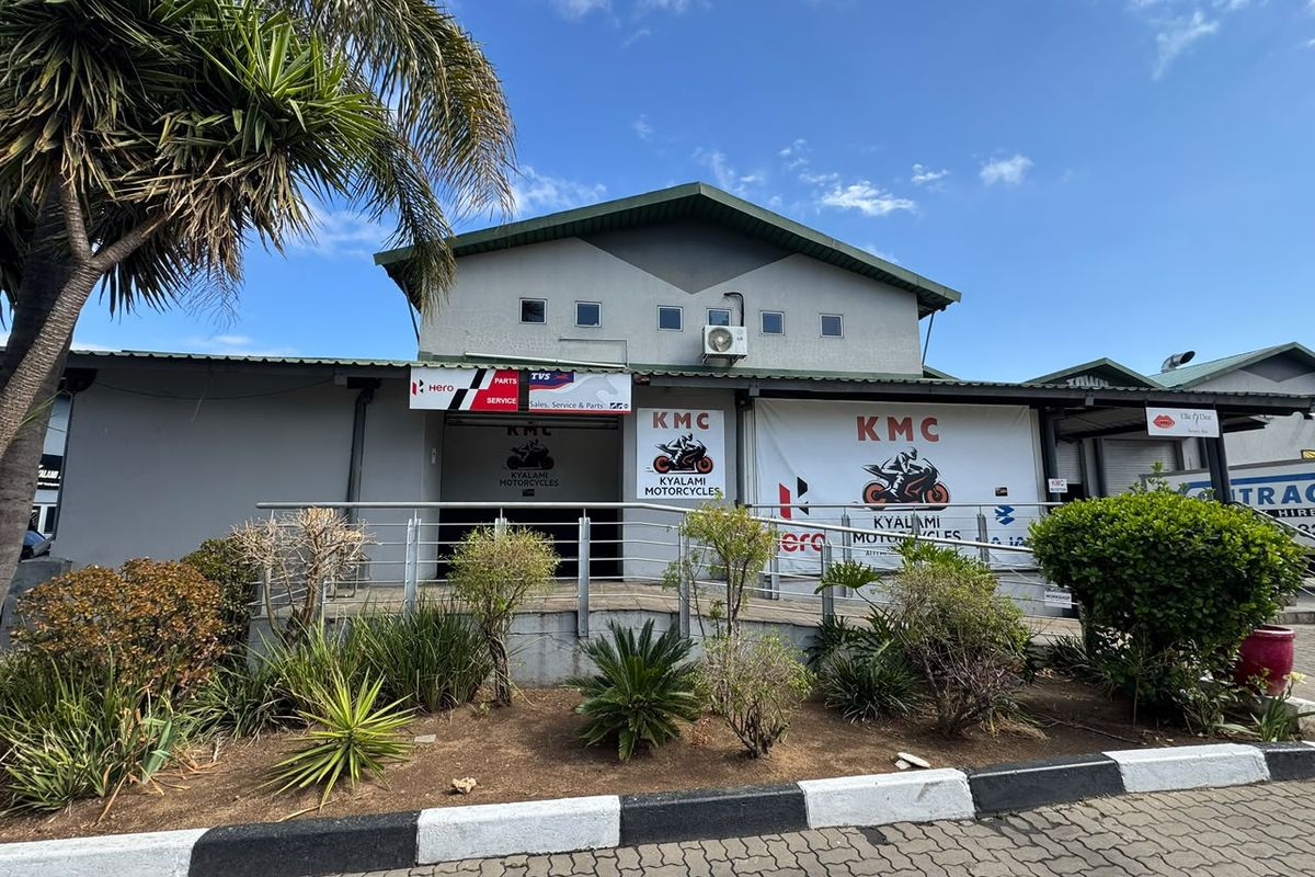 Front elevation of a commercial property featuring a metal roof, exterior air conditioning unit, and a paved entrance area with landscaped garden beds.