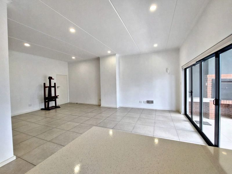 Spacious open-plan living area featuring neutral tiled flooring, recessed lighting, and large sliding glass doors leading to an outdoor patio with a built-in braai.