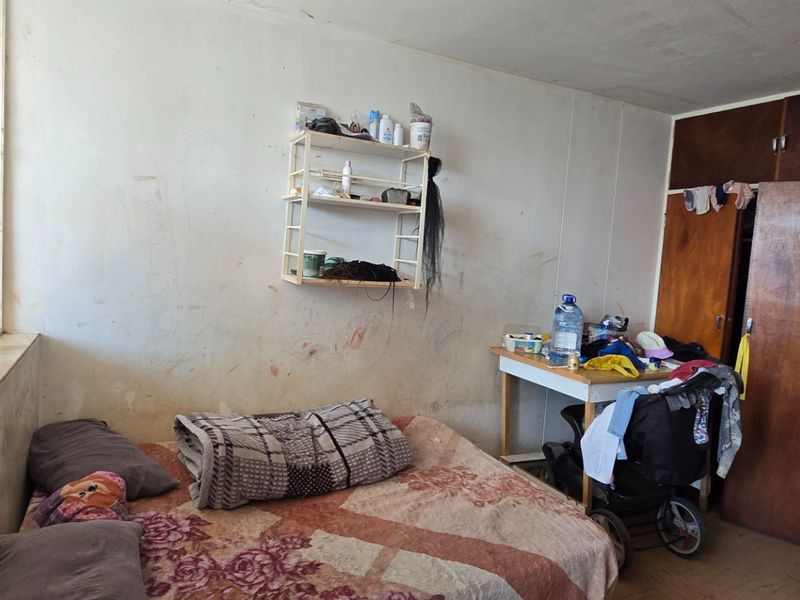 A bedroom featuring a built-in wooden wardrobe and a large window. The room requires cosmetic updates including wall repairs and fresh paint.