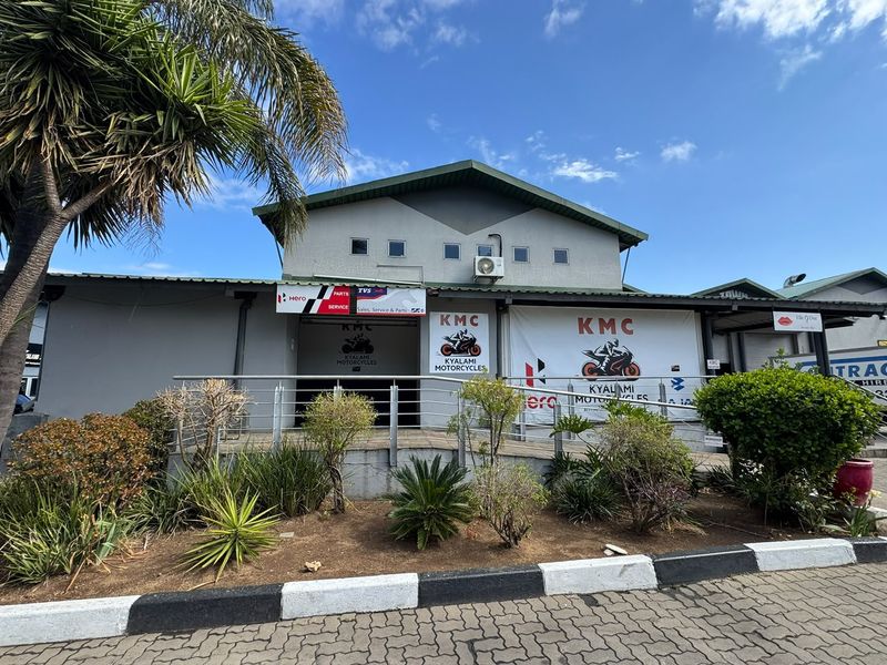Front elevation of a commercial property featuring a metal roof, exterior air conditioning unit, and a paved entrance area with landscaped garden beds.