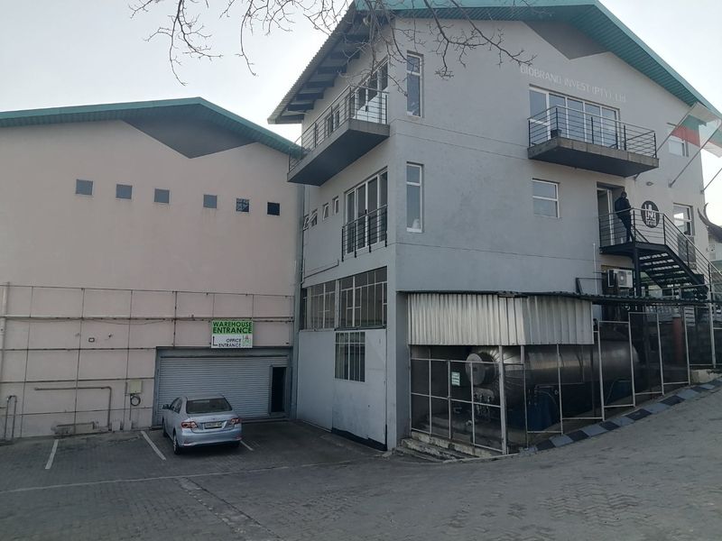 Commercial property exterior featuring a multi-level office building, warehouse entrance with roller shutter door, and an industrial storage tank in the parking area.