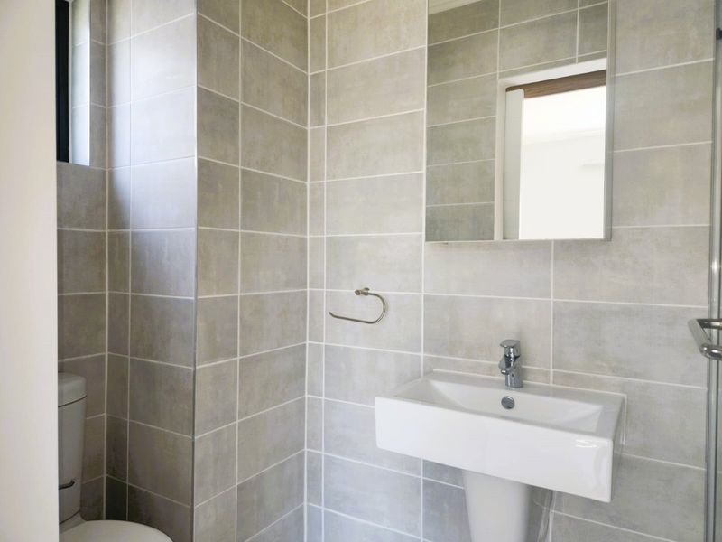 A clean, modern bathroom featuring neutral ceramic wall tiles, a pedestal sink, a wall-mounted mirror, and a shower enclosure.