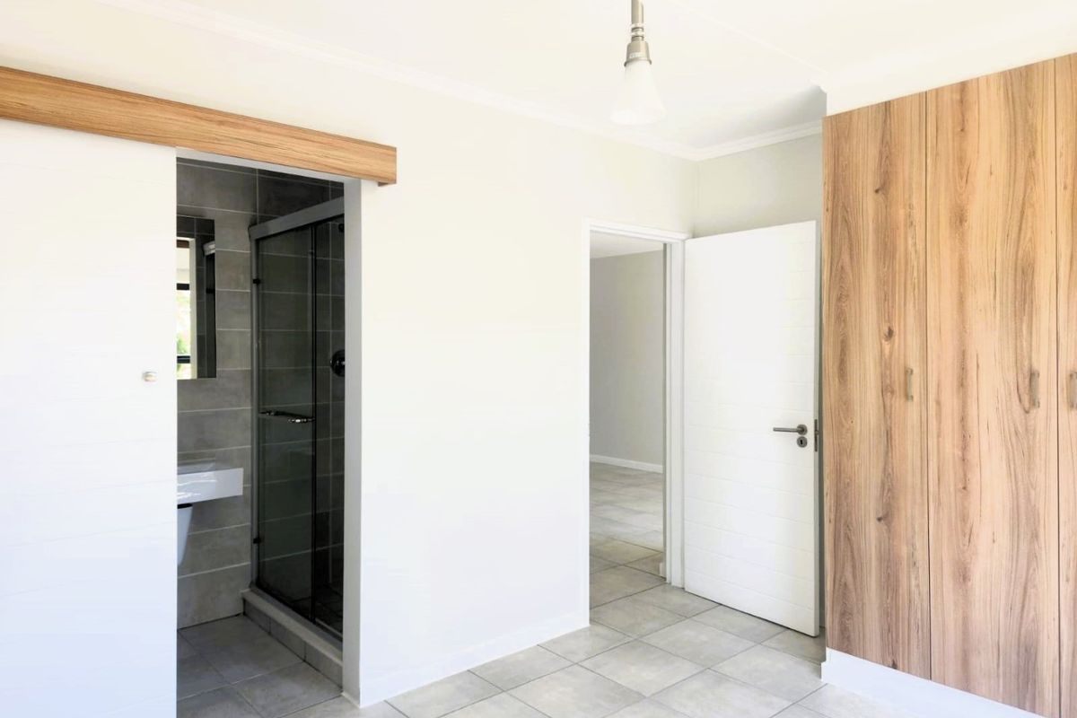 A bright, modern bedroom featuring neutral tiled flooring, built-in wood-finish cupboards, and a convenient en-suite bathroom with a glass shower enclosure.