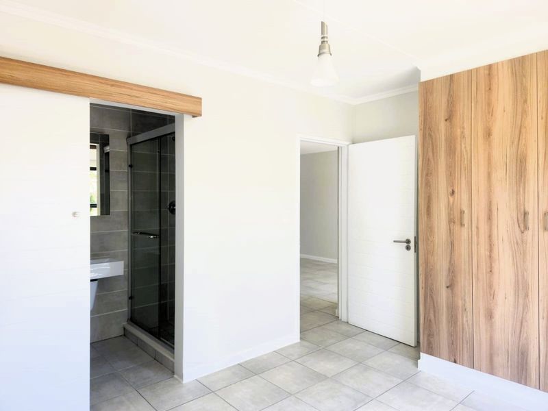 A bright, modern bedroom featuring neutral tiled flooring, built-in wood-finish cupboards, and a convenient en-suite bathroom with a glass shower enclosure.