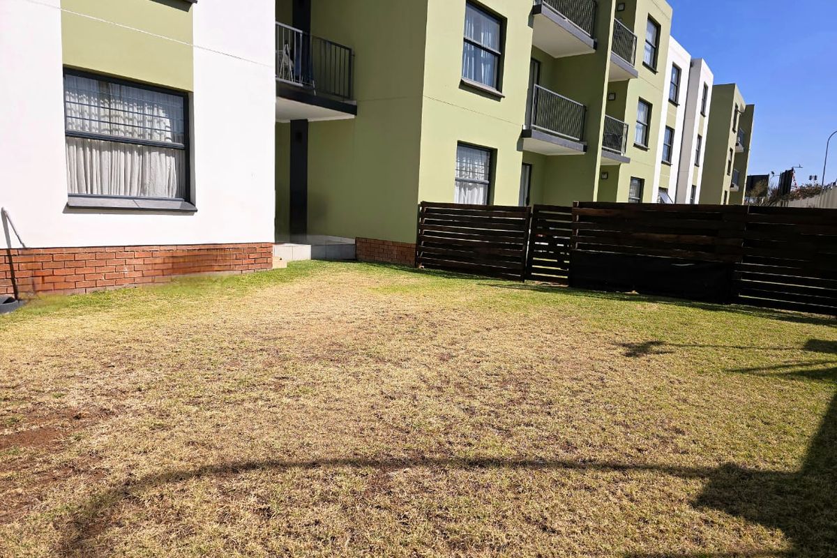 A rear view of an apartment complex featuring a private lawn area, timber fencing, and multi-level balconies with brick and rendered facade finishes.