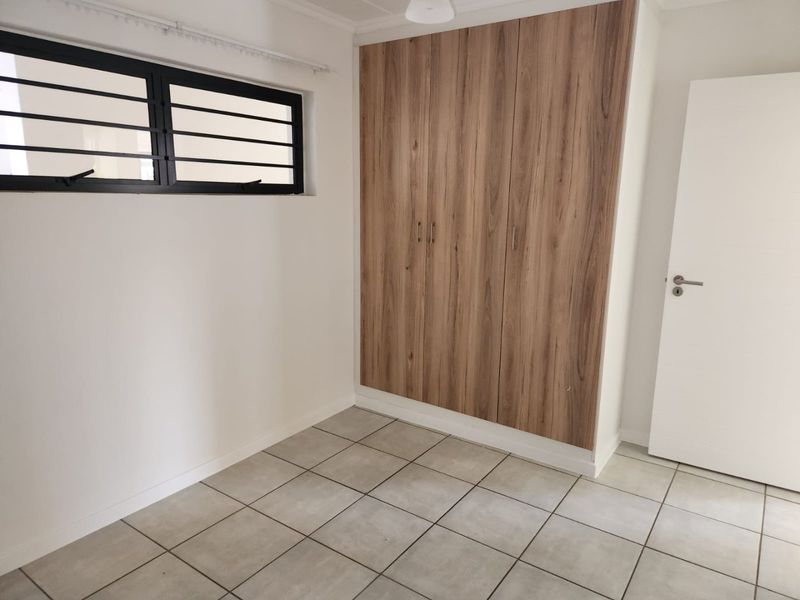 A clean, empty bedroom featuring modern wood-finish built-in cupboards, neutral tiled flooring, and a window with security bars.