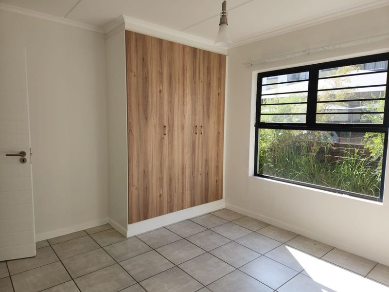 A bright, empty bedroom featuring modern wood-finish built-in cupboards, neutral tiled flooring, and a large window with security bars.