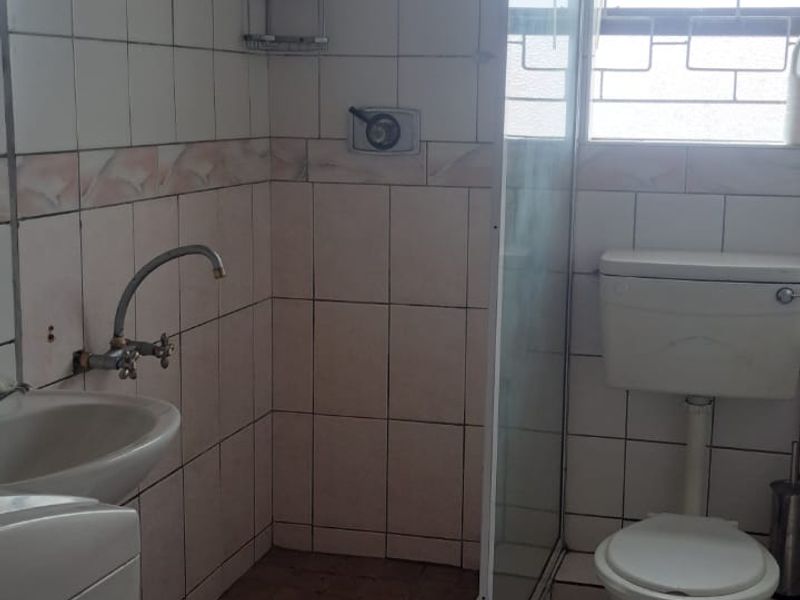 A functional bathroom featuring a shower enclosure, toilet, and wash basin with tiled finishes and security bars on the window.