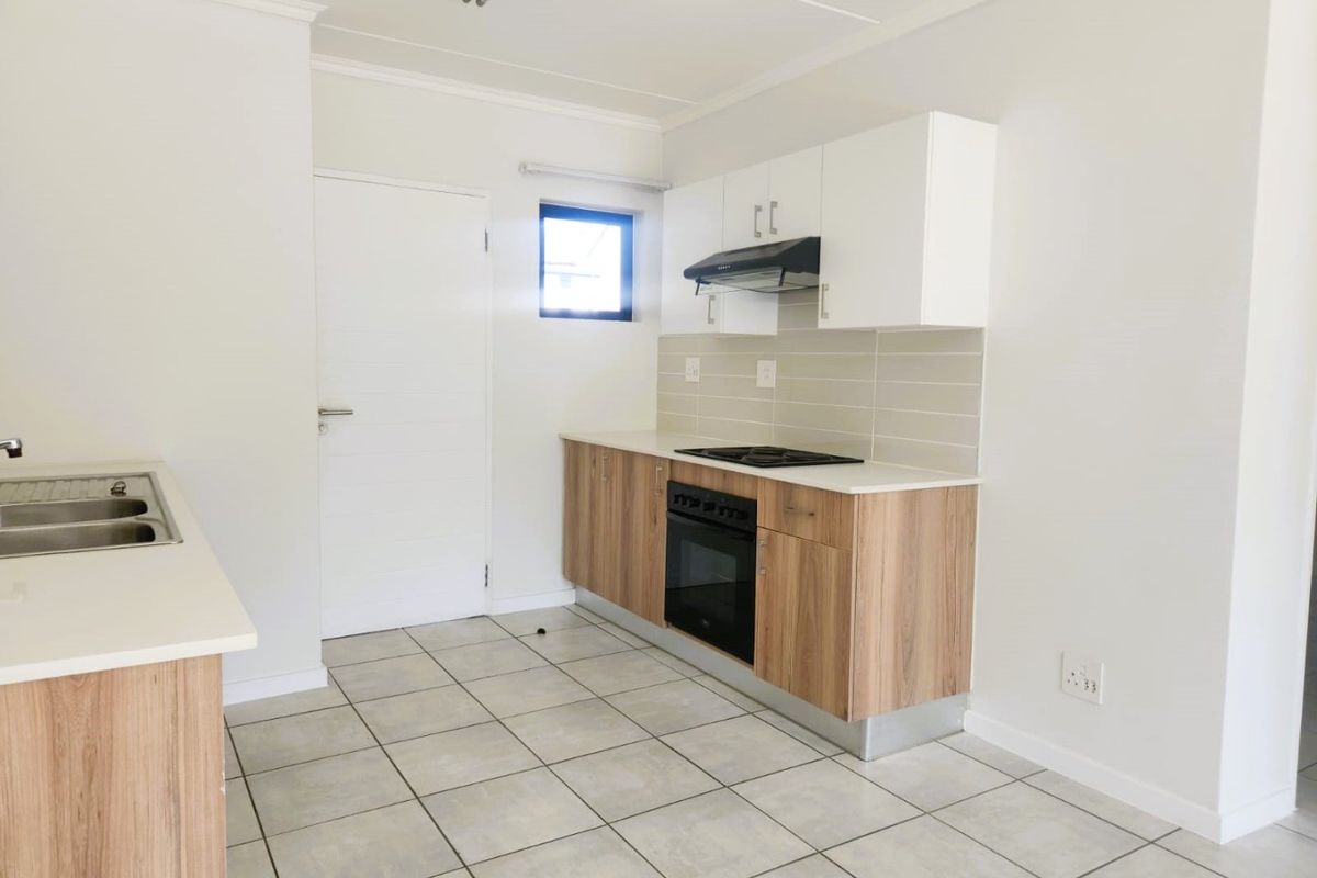 A clean, modern kitchen featuring light wood-finish cabinetry, stone countertops, a built-in oven with hob, and neutral tiled flooring.