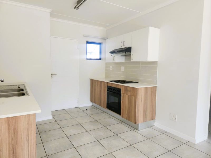 A clean, modern kitchen featuring light wood-finish cabinetry, stone countertops, a built-in oven with hob, and neutral tiled flooring.