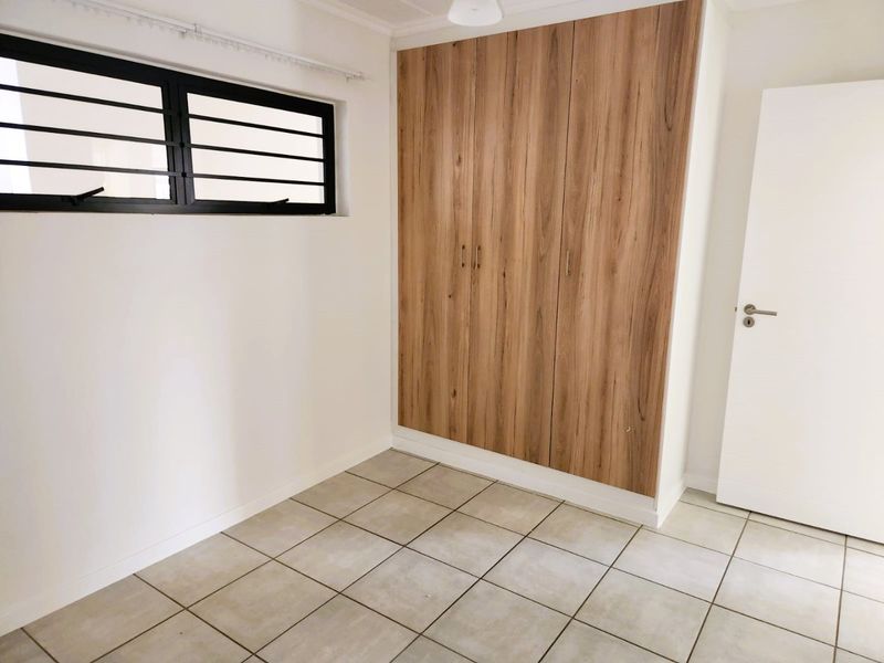 A clean, empty bedroom featuring modern wood-finish built-in cupboards, neutral floor tiles, and a window with security bars.