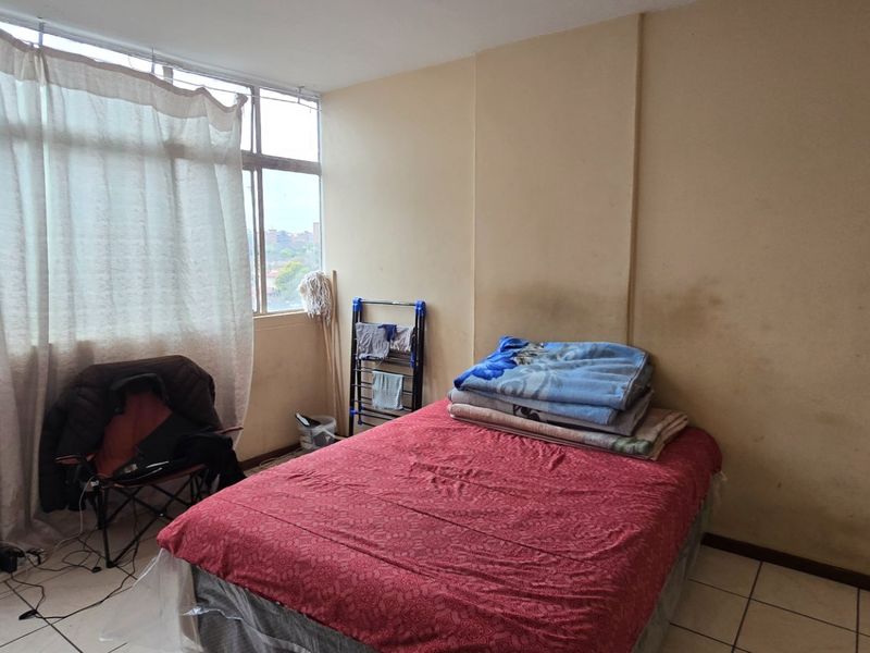 A bedroom featuring tiled flooring, a built-in wardrobe, and a large window. The walls show signs of staining and require maintenance.