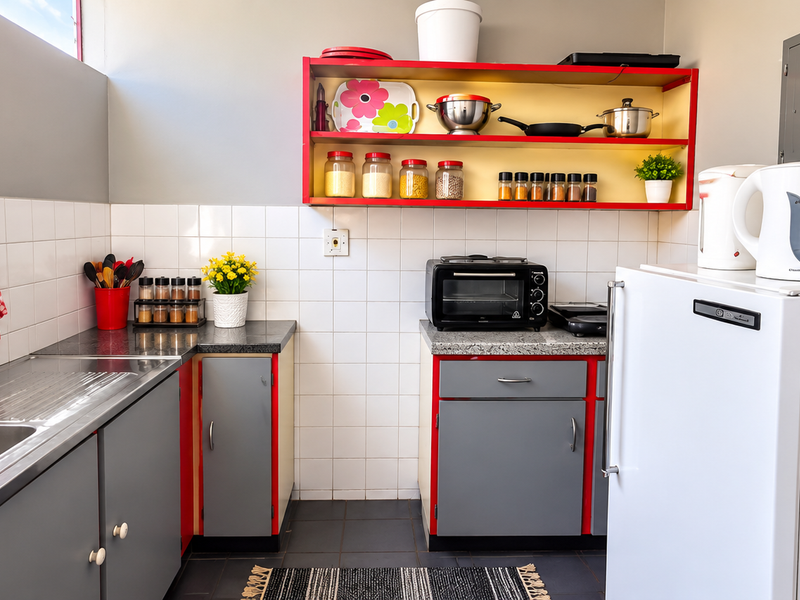A functional kitchen featuring grey cabinetry with red accents, granite countertops, a stainless steel sink, and a tiled backsplash.