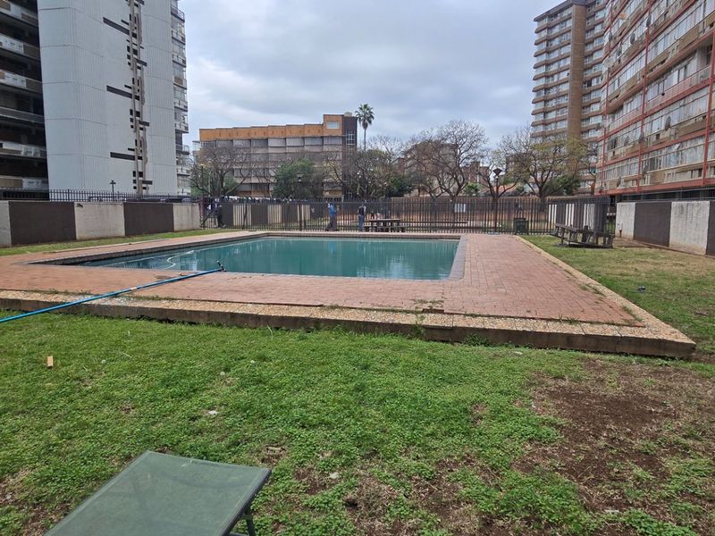 Communal swimming pool area featuring brick coping and perimeter fencing, situated within an apartment complex with surrounding lawn and boundary walls.