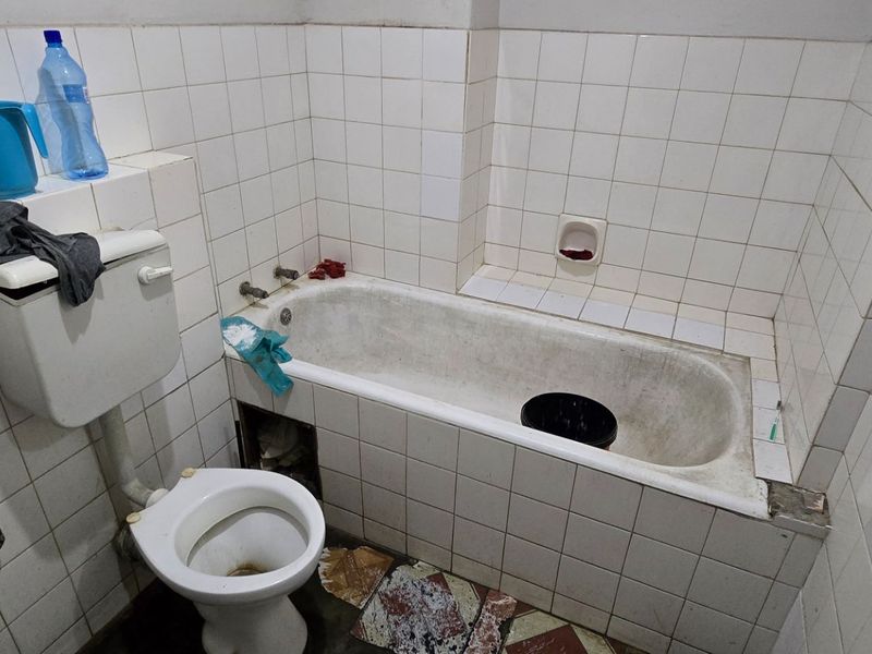 A dated bathroom featuring a standard bathtub, toilet, and washbasin with white wall tiling, requiring significant renovation and modernization of fixtures and finishes.