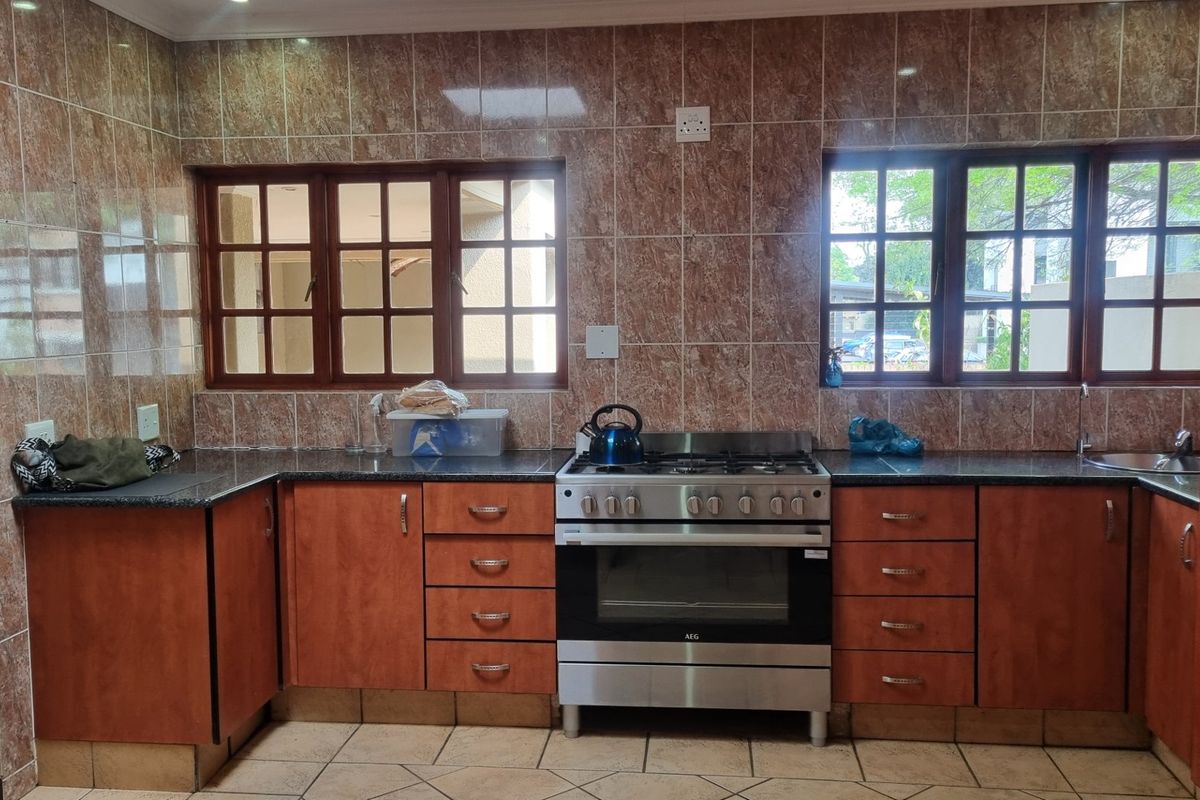 A functional kitchen featuring wooden cabinetry, granite countertops, a gas stove with oven, and tiled wall finishes with natural light from timber-framed windows.