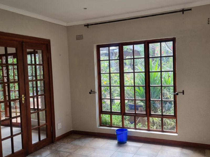 A bright, empty living area featuring classic wooden french doors and large paneled windows overlooking the garden, finished with neutral walls and tiled flooring.