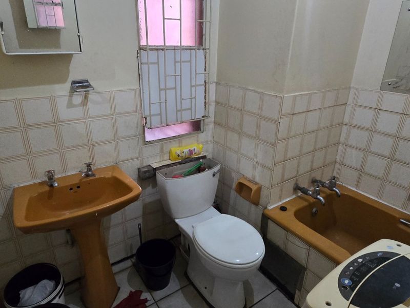 A dated bathroom featuring original colored sanitaryware, ceramic wall tiling, and a window with security bars, requiring modernization and renovation.