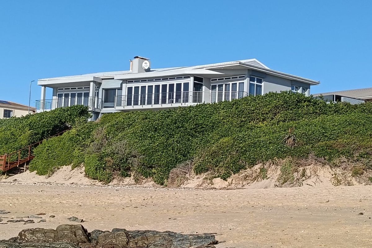 A view of the rear elevation of a coastal house, featuring large windows, a balcony, and direct access to the beach via a staircase.