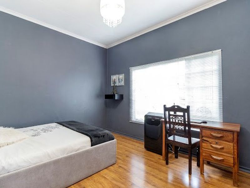 A well-maintained bedroom featuring dark grey walls, laminate flooring, and a large window with venetian blinds, complemented by a decorative crystal chandelier.