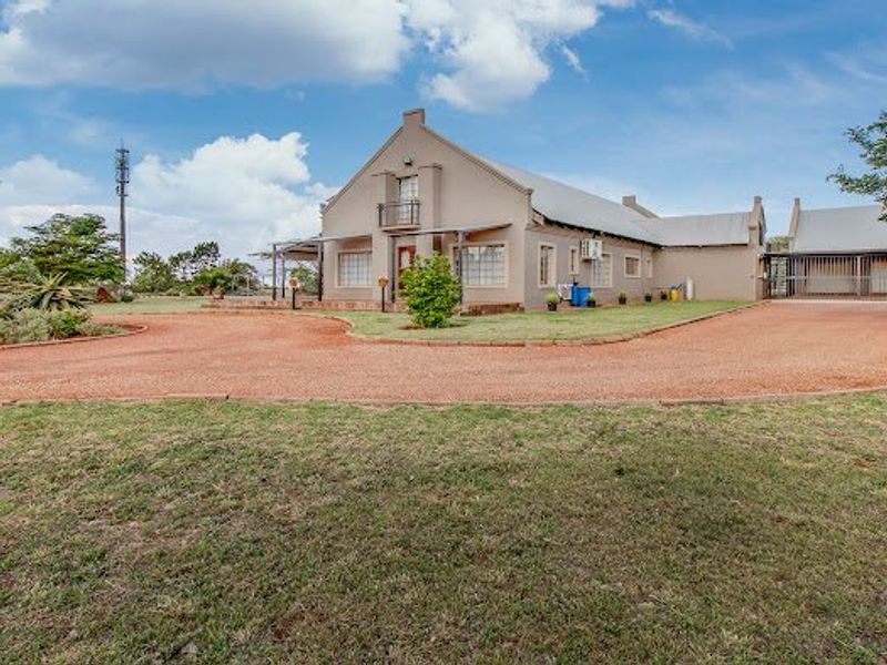 A well-maintained gabled house featuring a gravel driveway, landscaped garden, and an upper-level balcony, set within a spacious rural or estate environment.