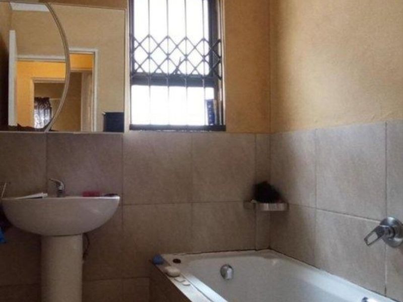 A functional bathroom featuring a pedestal basin, bathtub with wall-mounted mixer, and tiled wall finishes with a security-gated window.