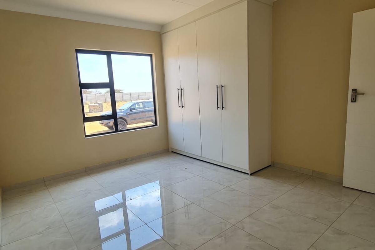 A clean, empty bedroom featuring modern tiled flooring, built-in cupboards, and an aluminium-framed window providing natural light.