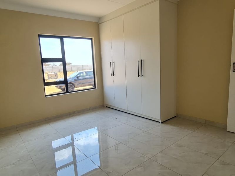 A clean, empty bedroom featuring modern tiled flooring, built-in cupboards, and an aluminium-framed window providing natural light.