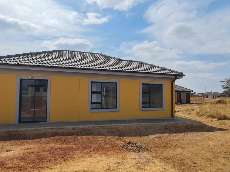 A modern single-story house exterior featuring yellow painted walls, a tiled roof, and aluminium-framed windows, set on a developing residential plot.