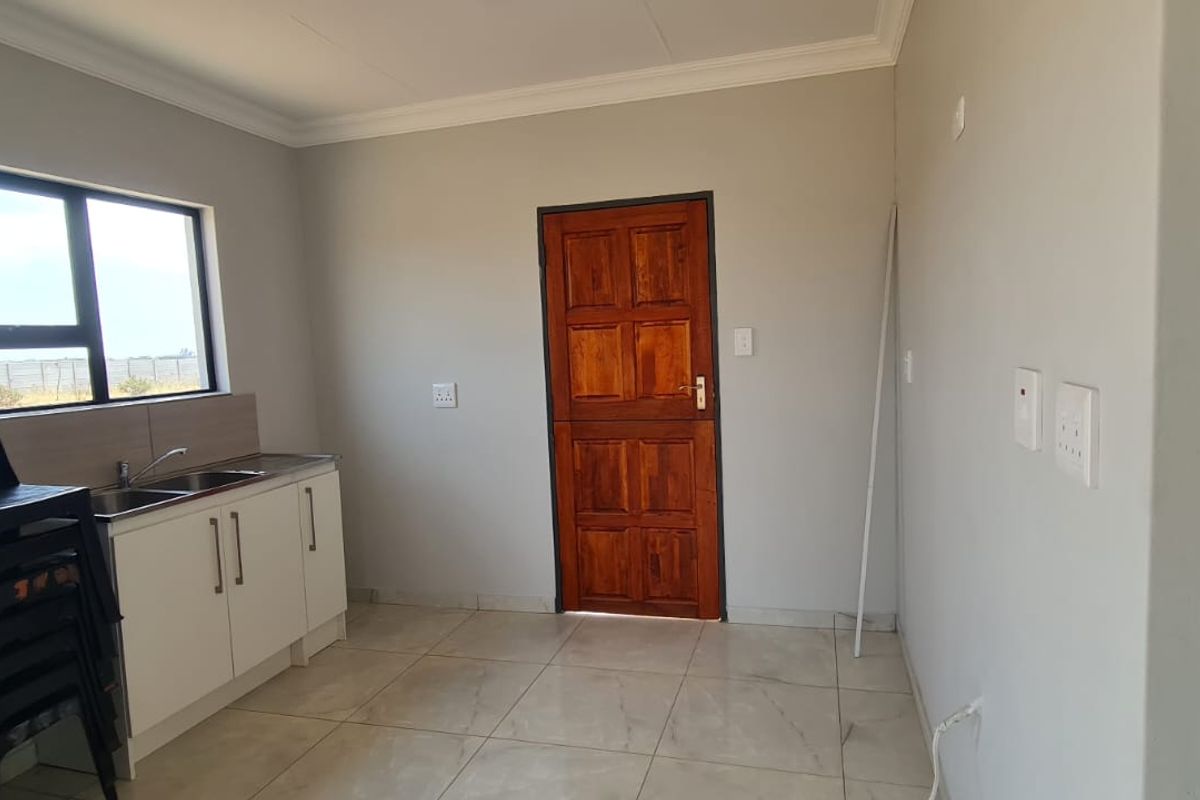 A clean, empty kitchen area featuring tiled flooring, white cabinetry with a stainless steel sink, and a solid timber door.