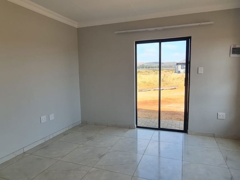 A bright, empty living area featuring neutral tiled flooring, modern aluminium window frames, and a sliding glass door leading to the exterior.