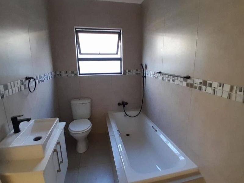 A clean, modern bathroom featuring a built-in bathtub, a contemporary vanity with basin, and a toilet, finished with neutral wall and floor tiles.