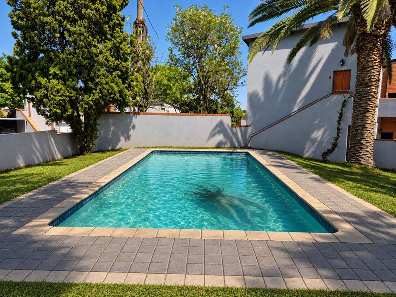 A well-maintained outdoor swimming pool area featuring a paved surround, landscaped garden, and boundary wall within an apartment complex setting.
