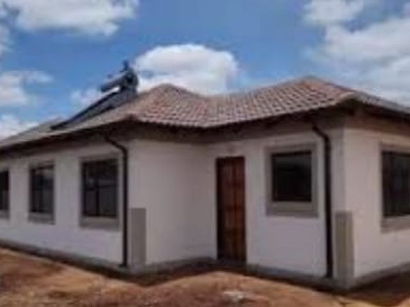 A single-story residential house featuring a tiled roof, solar water heating system, and exterior plaster finish, set on an undeveloped plot.