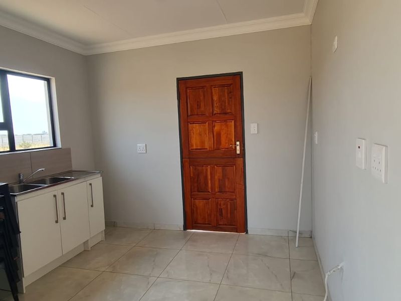 A clean, empty kitchen area featuring tiled flooring, white cabinetry with a stainless steel sink, and a solid timber door.