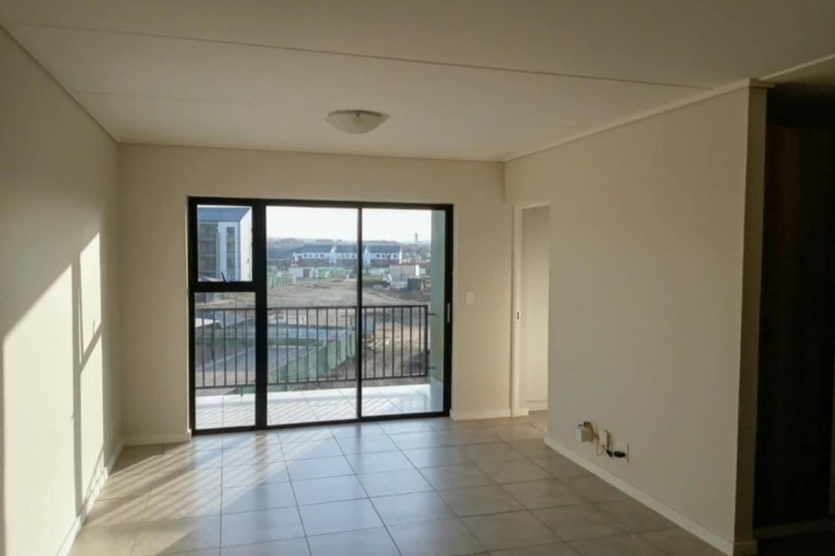 An empty, open-plan living area featuring neutral tiled flooring, a ceiling light, and a sliding glass door leading to a balcony.